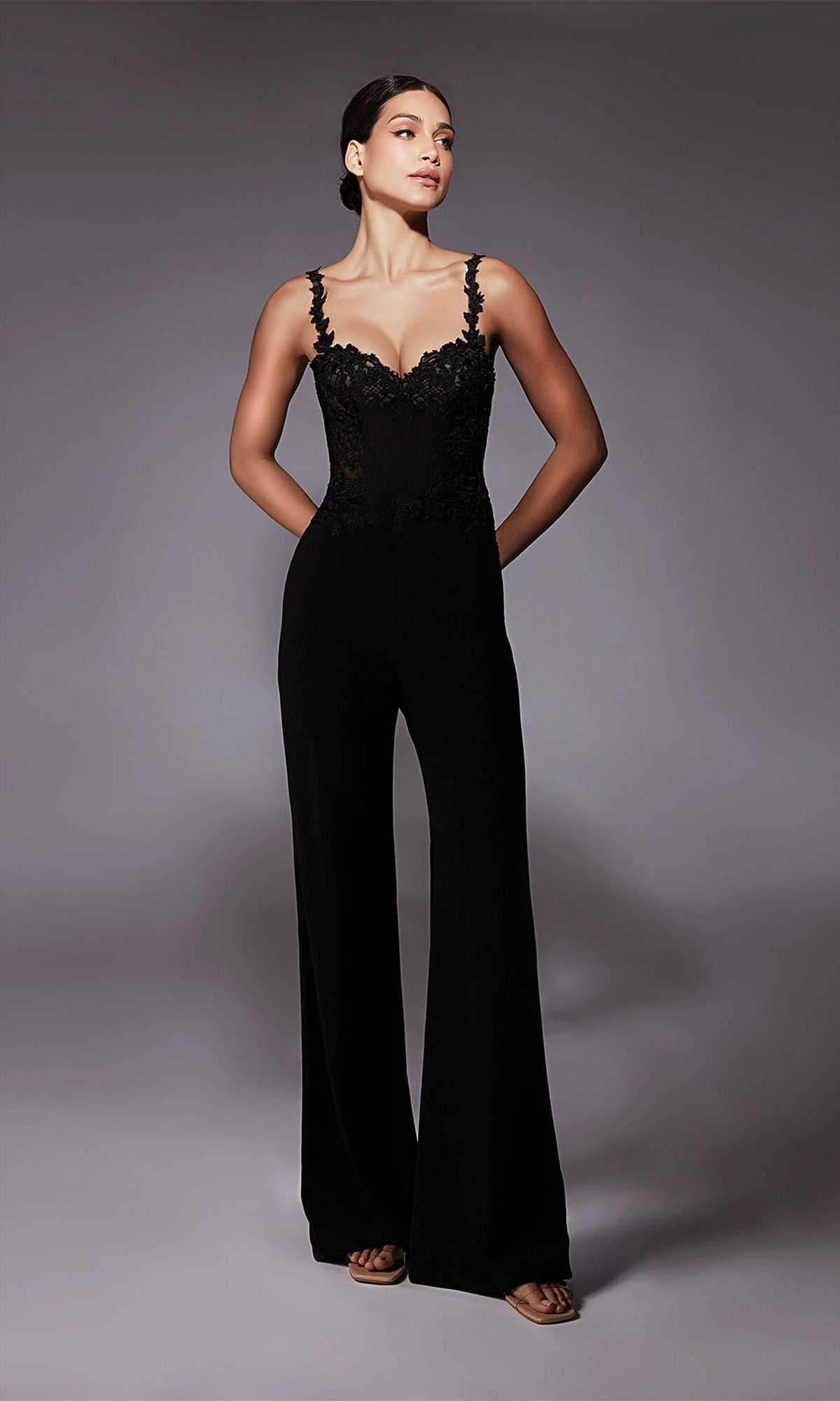 A woman poses in a studio wearing the Long Formal Jumpsuit: Alyce 70074 in black, featuring wide-leg pants, lace trim, and spaghetti straps, with her arms relaxed at her sides.